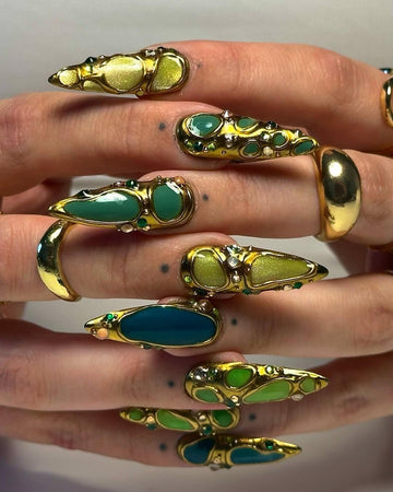 Handmade press-on nails with gold, green, and blue abstract designs, worn with gold rings.