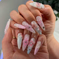 Hand with long, stiletto press-on nails featuring 3D pink floral designs and rhinestone accents.