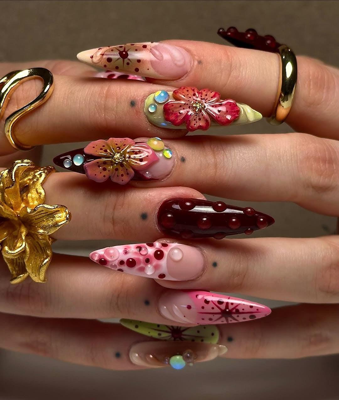 Hand with long stiletto press-on nails featuring 3D floral designs, rhinestones, and gold rings
