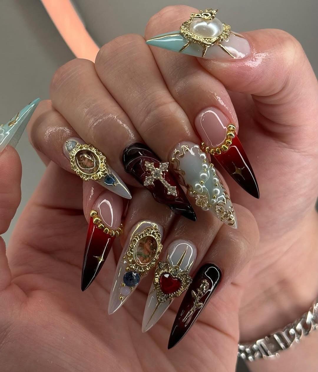 Hand holding custom press-on nails with ornate gold designs, gems, and heart embellishments.