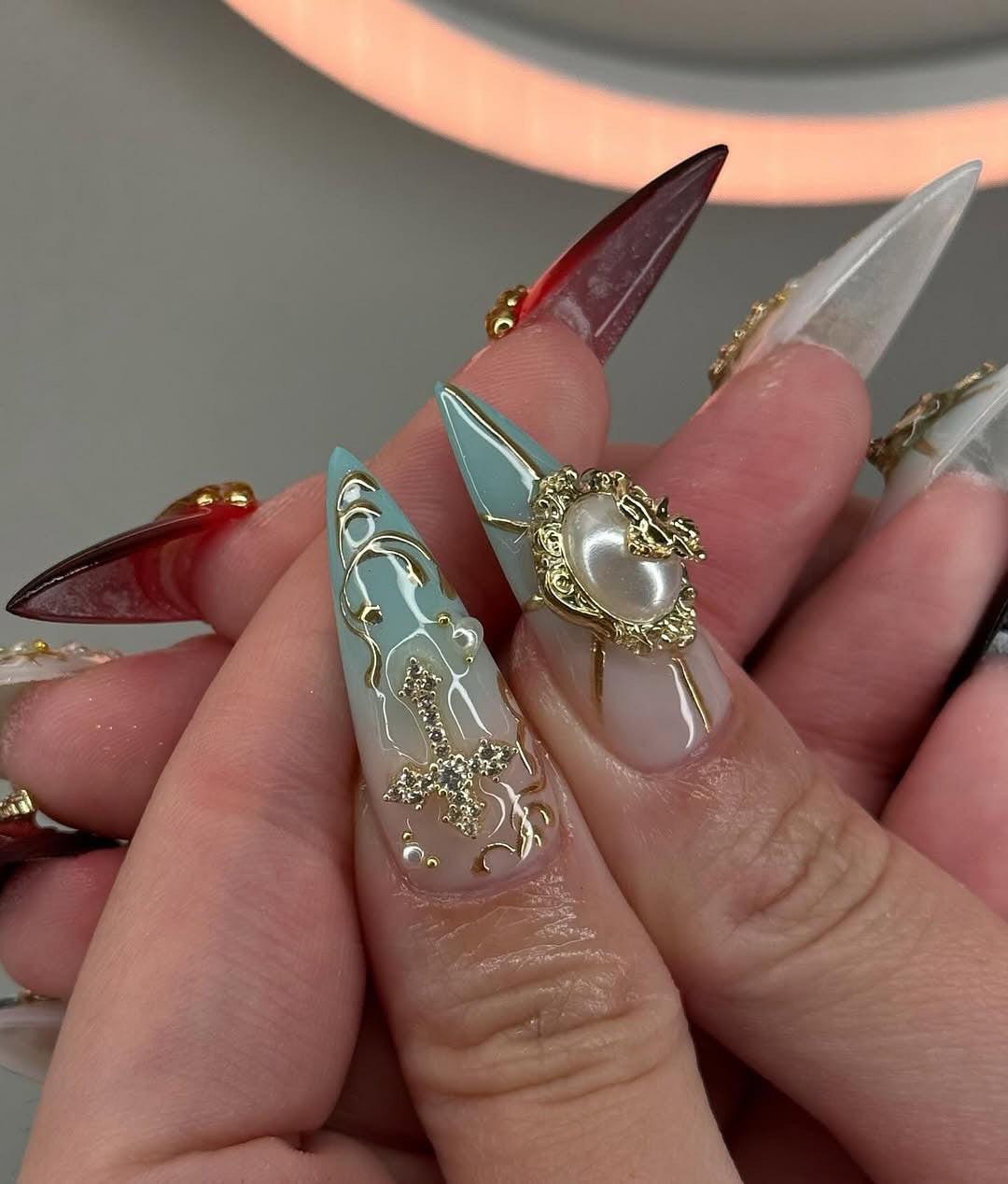 Hand holding custom press-on nails with ornate gold designs, gems, and heart embellishments.