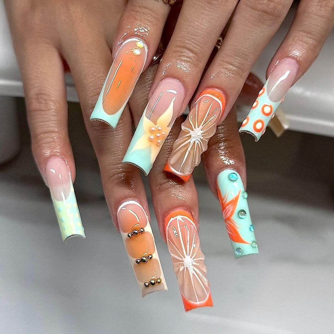 Hand with long press-on nails in orange, turquoise, and nude, featuring 3D floral and geometric designs