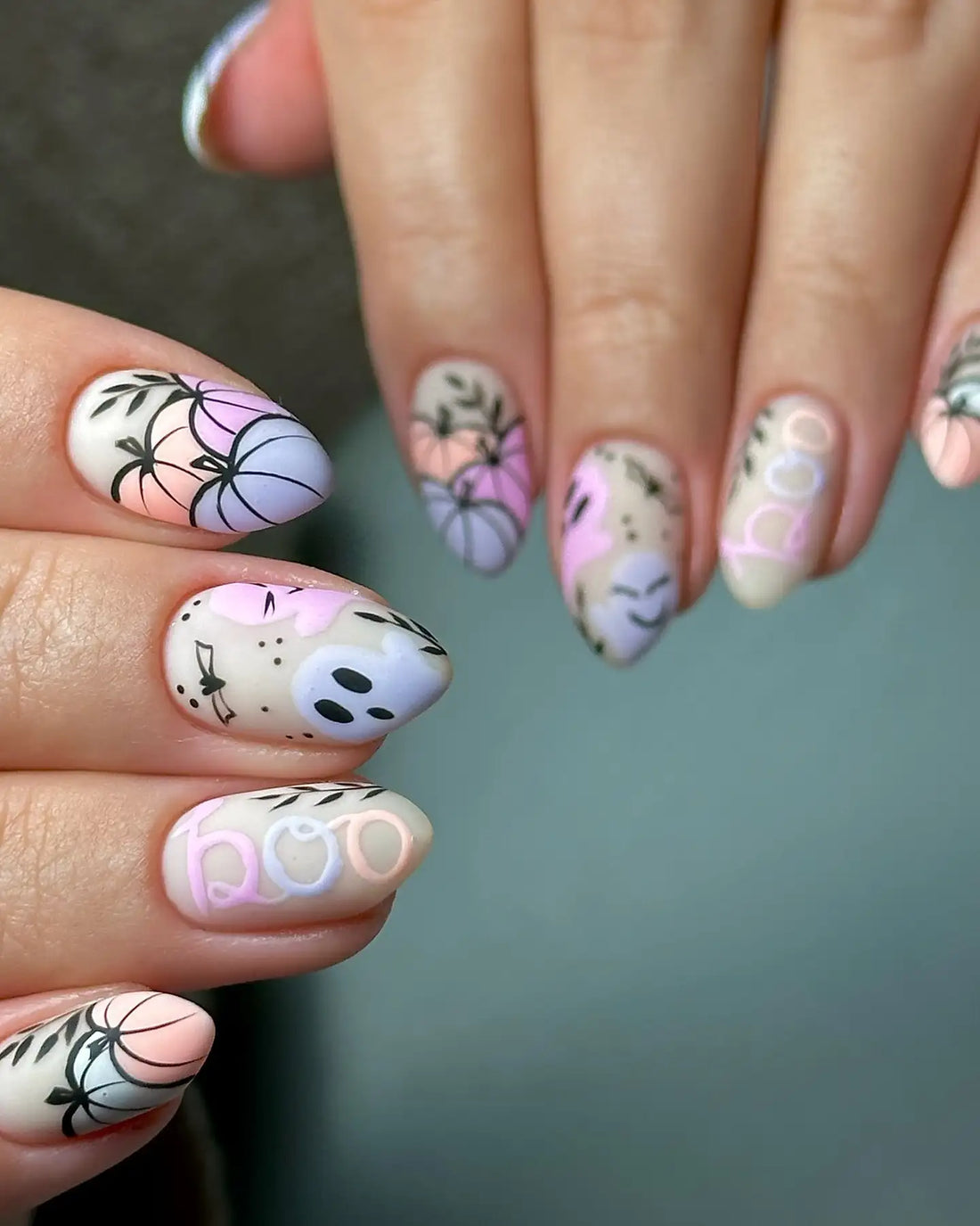 Hand with pastel Halloween press-on nails featuring pumpkins, ghosts, boo text, and leafy designs.
