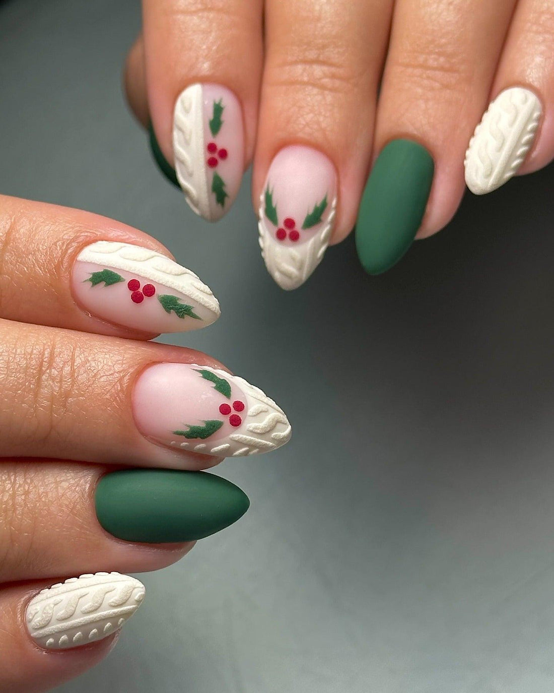 Hand with almond-shaped press-on nails featuring matte green, cable-knit texture, and holly berry holiday nail art.