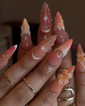 Hand with long, almond-shaped press-on nails featuring pink ombre and 3D floral nail art.