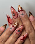 Hand with long, almond-shaped press-on nails featuring cherry art, red designs, gold bows, and brown marble tips.
