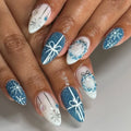 Hand with blue and white press-on nails featuring snowflakes, bows, and winter designs.