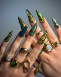 Hand with long, sculpted green and gold gemstone press-on nails, gold rings, elegant manicure