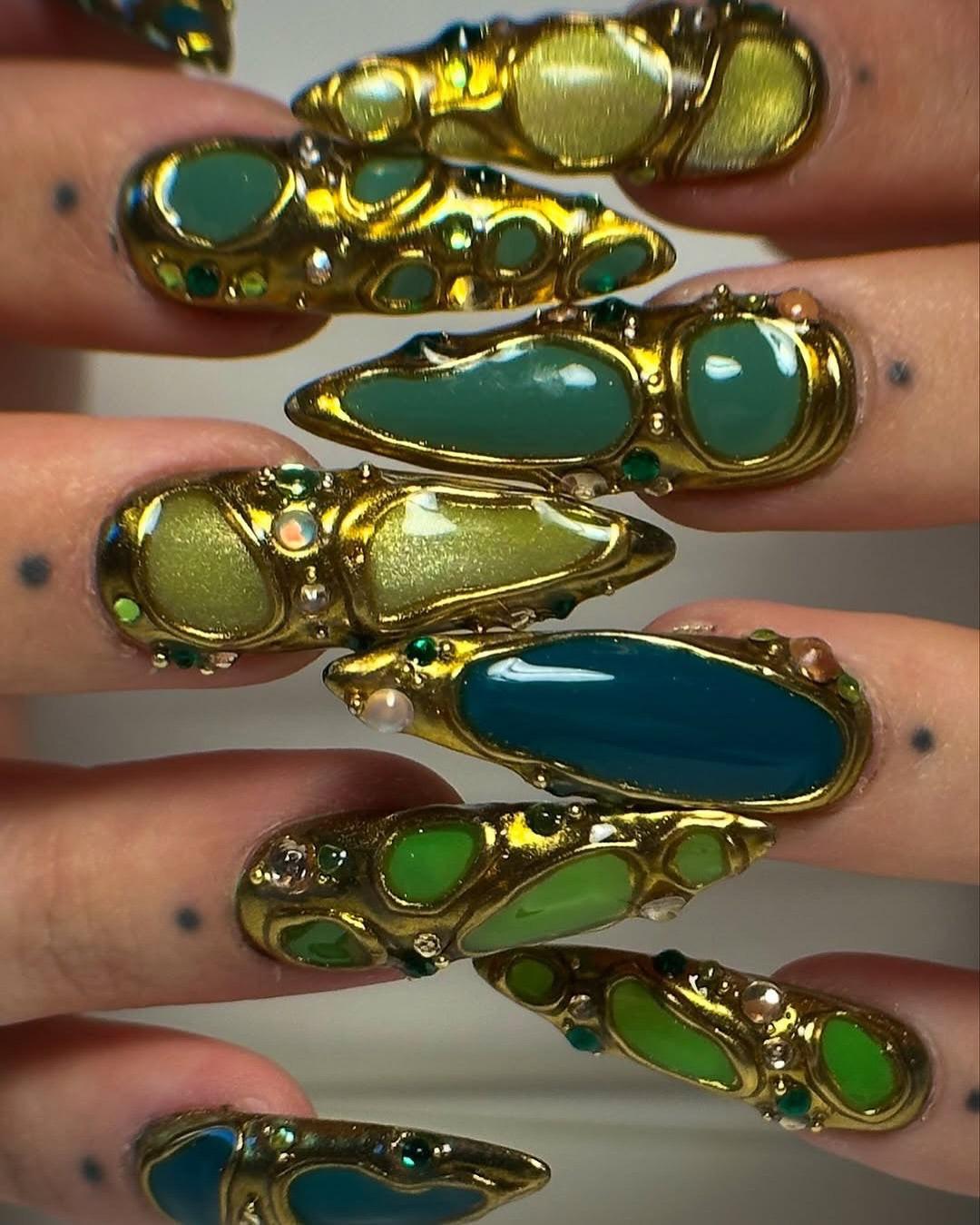 Hand with long press-on nails featuring gold metallic accents and green gemstone designs