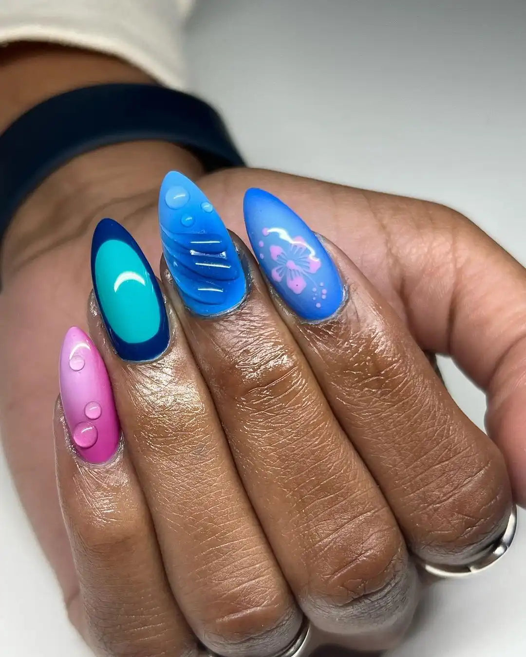Hand with vibrant handmade press-on nails in blue, teal, and pink, featuring hibiscus and water droplet designs.