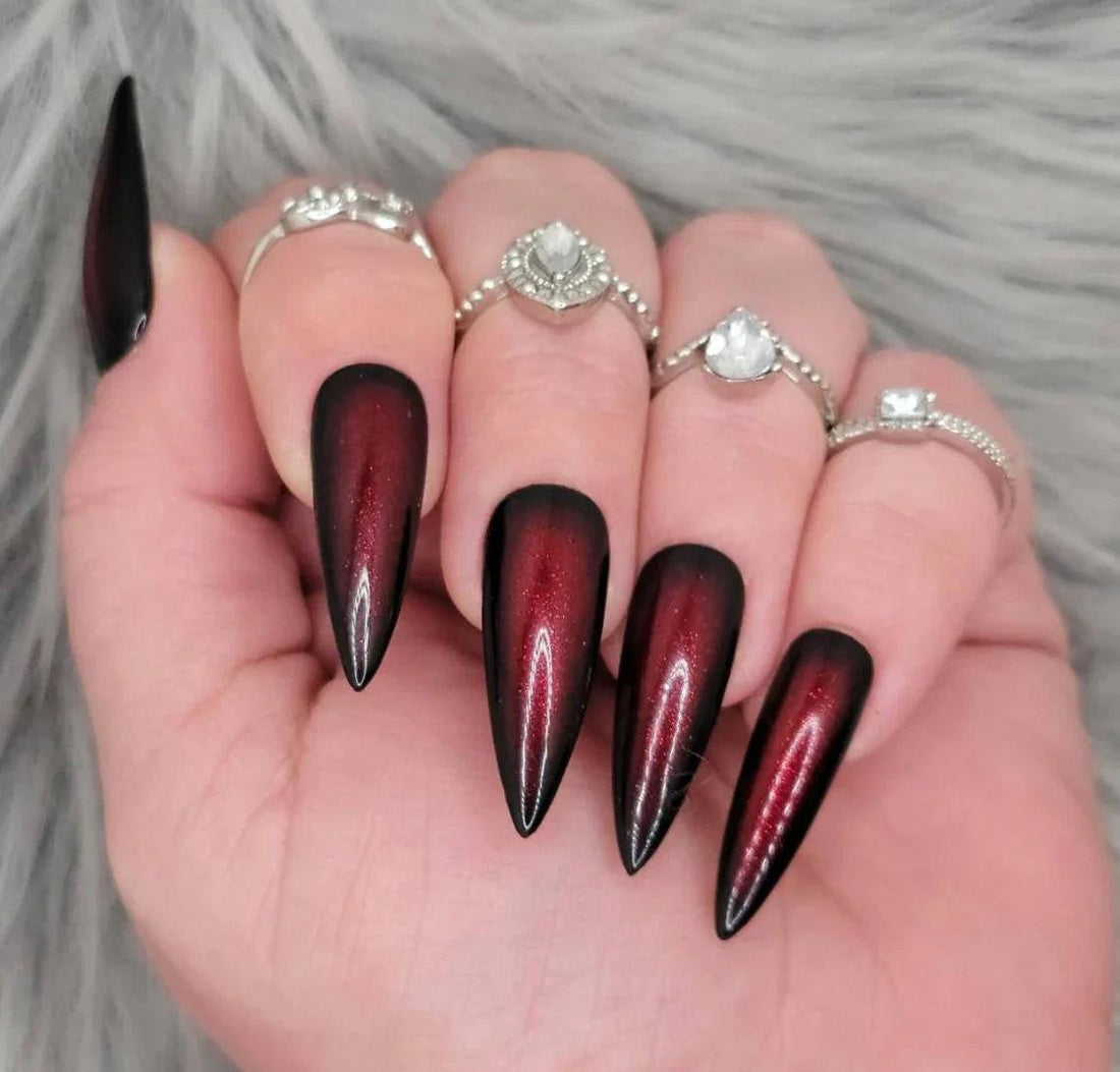 Long stiletto black and red ombre press-on nails with gothic style, worn with silver rings