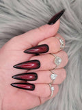 Long stiletto press-on nails in glossy black and red ombre, shown with silver rings.