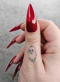 Hand with long, glossy stiletto red press-on nails and a small ghost tattoo on the thumb.