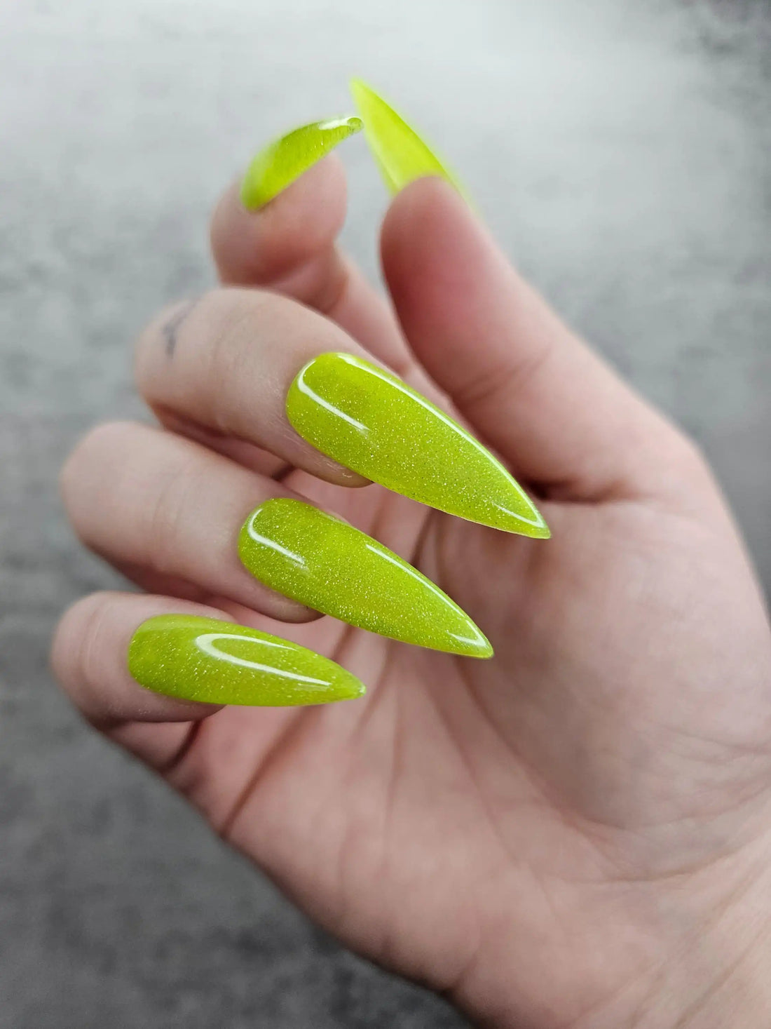 Hand with long green glitter press-on nails, almond shape, on a neutral background.