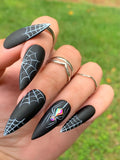Black matte stiletto press-on nails with white spider web designs and rhinestone spider accent