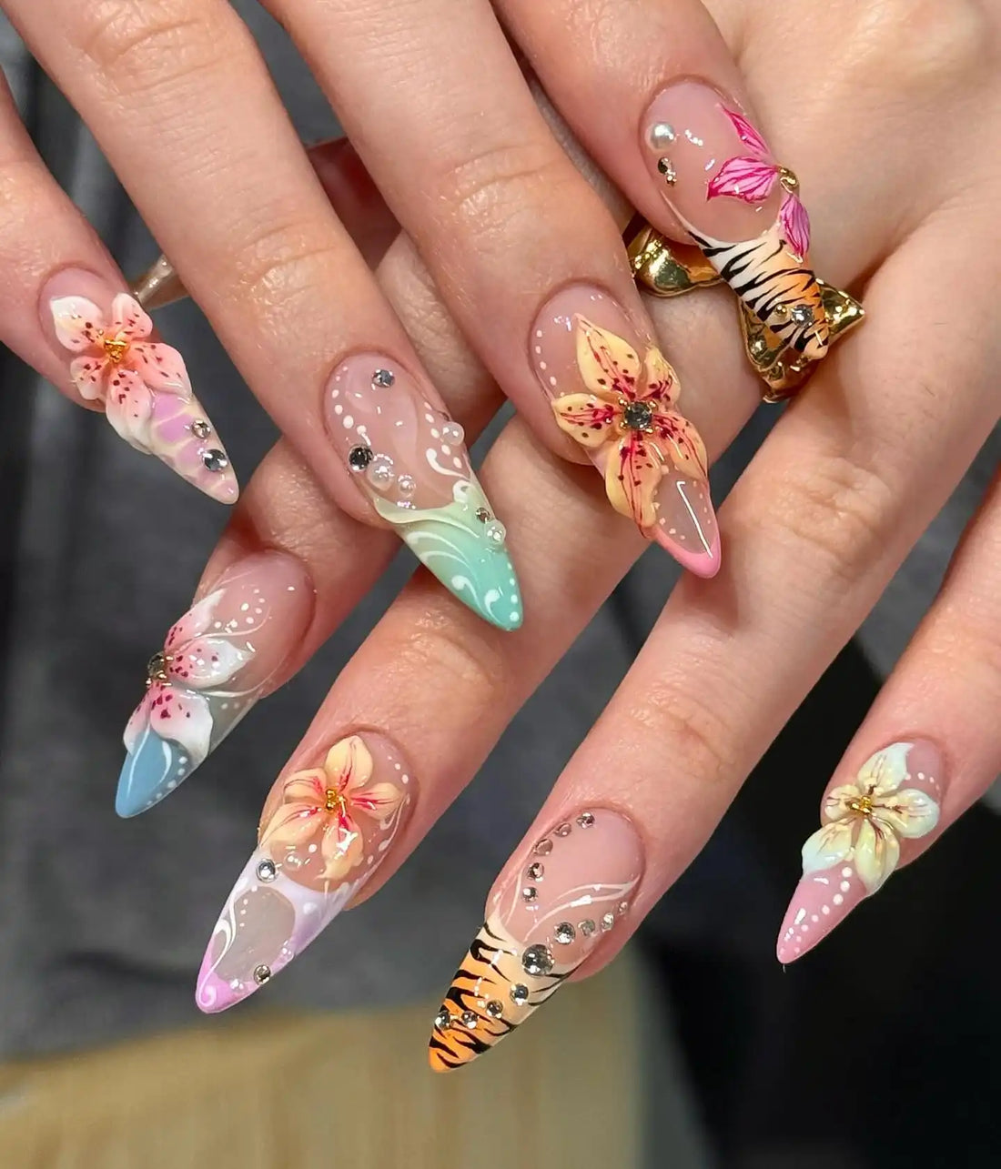 Hand holding long stiletto press-on nails with 3D floral designs, ombre colors, tiger print, and rhinestones