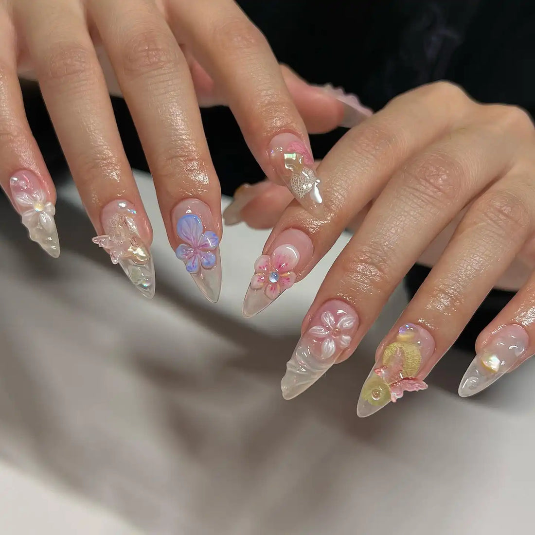 Hand model with long, almond-shaped press-on nails featuring 3D floral and butterfly designs in pastel colors.
