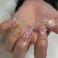 Hand with long press-on nails featuring 3D floral and butterfly spring nail art designs.