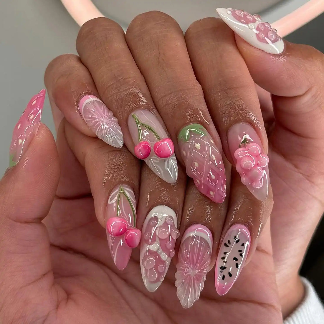 Hand with long pink press-on nails featuring 3D fruit designs including strawberries and cherries