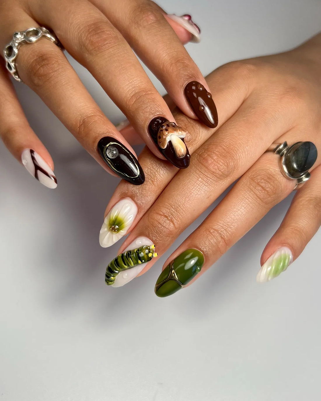 Hand with handmade 3D press-on nails featuring mushroom, caterpillar, and forest designs in green and brown.