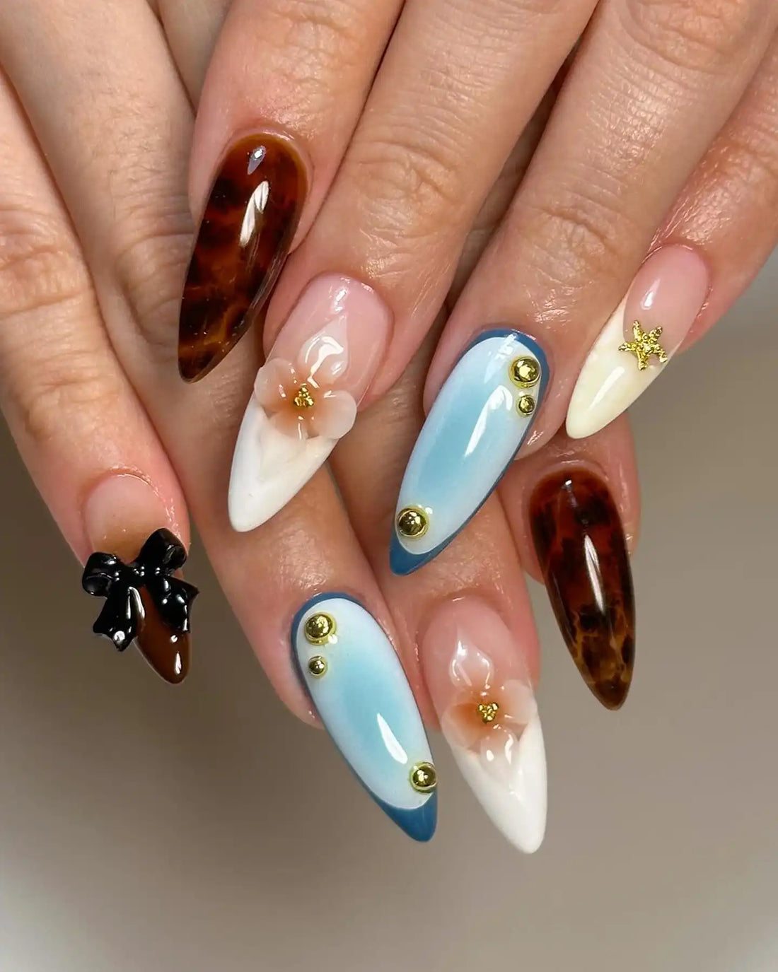 Hand with long stiletto press-on nails featuring brown, blue, and white designs, 3D flowers, gold studs, and a black bow.