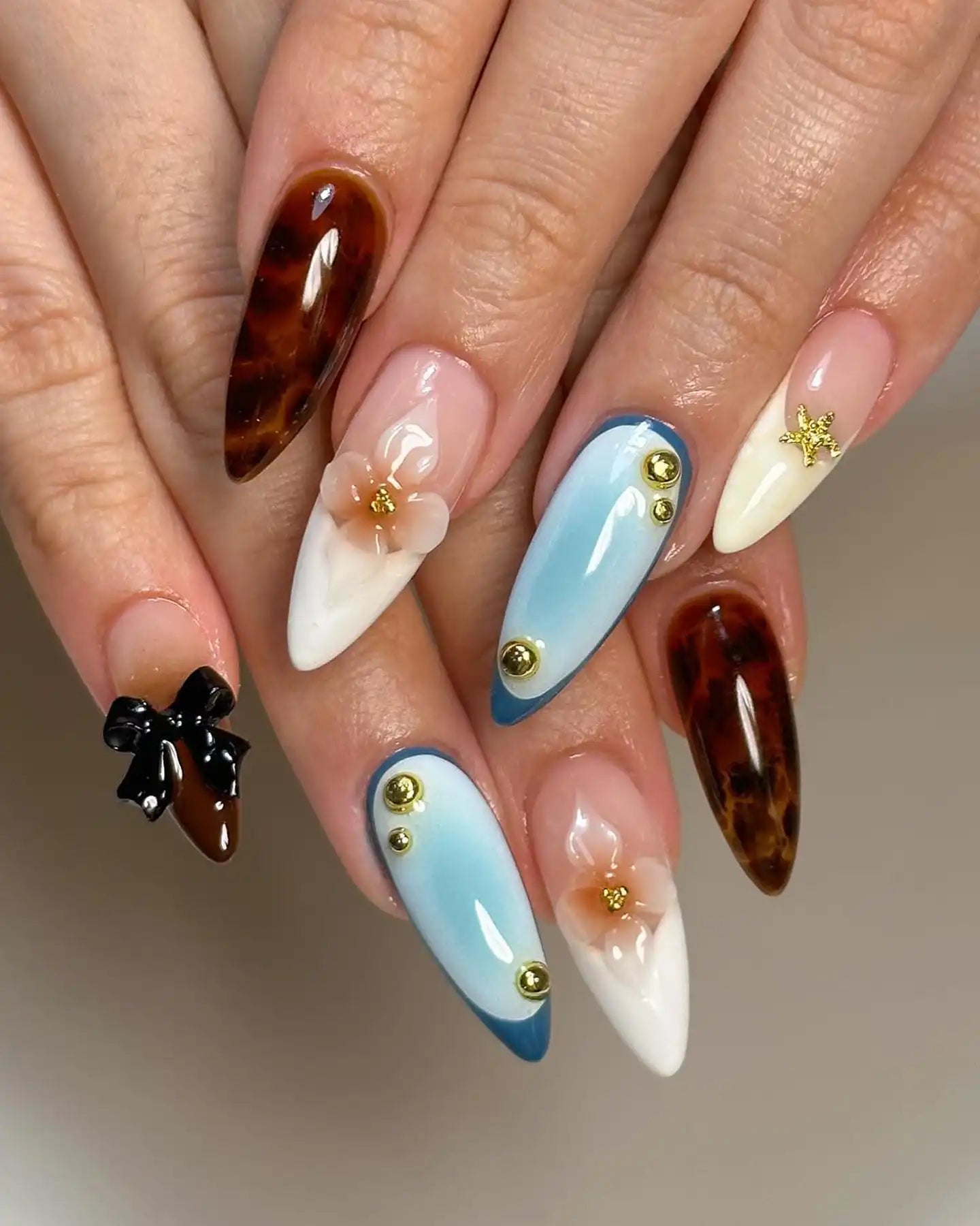 Hand with long stiletto press-on nails featuring brown, blue, and white designs, 3D flowers, gold studs, and a black bow.