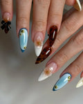 Handmade press-on nails with blue gradients, 3D floral accents, gold studs, and a black ribbon design.