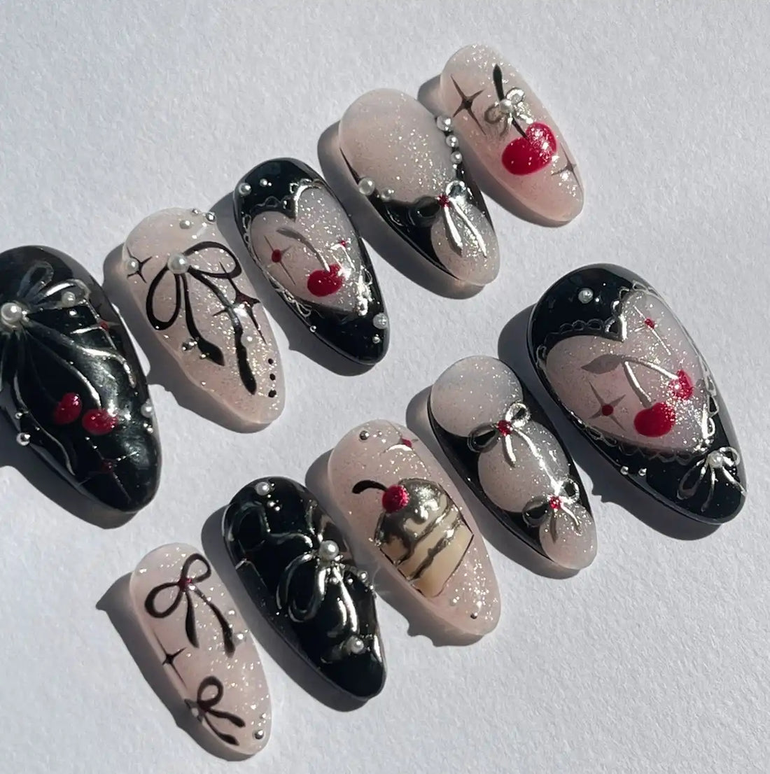 Handmade press-on nails with black and pink designs, cherries, bows, pearls, and glitter accents.
