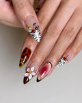 Hand with handmade press-on nails featuring zebra and tortoiseshell patterns, cherries, flowers, and gold accents.