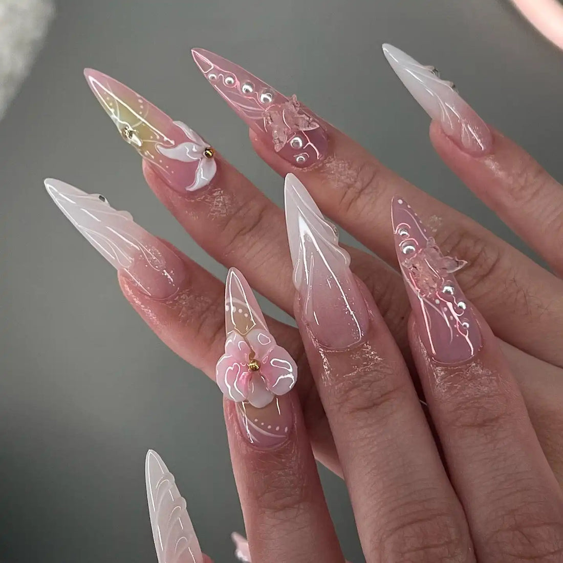 Long stiletto press-on nails with 3D flowers, butterfly accents, pearls, and pastel designs.