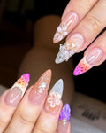 Hand with handmade press-on nails featuring 3D butterfly and flower designs, pastel colors, and gem accents.