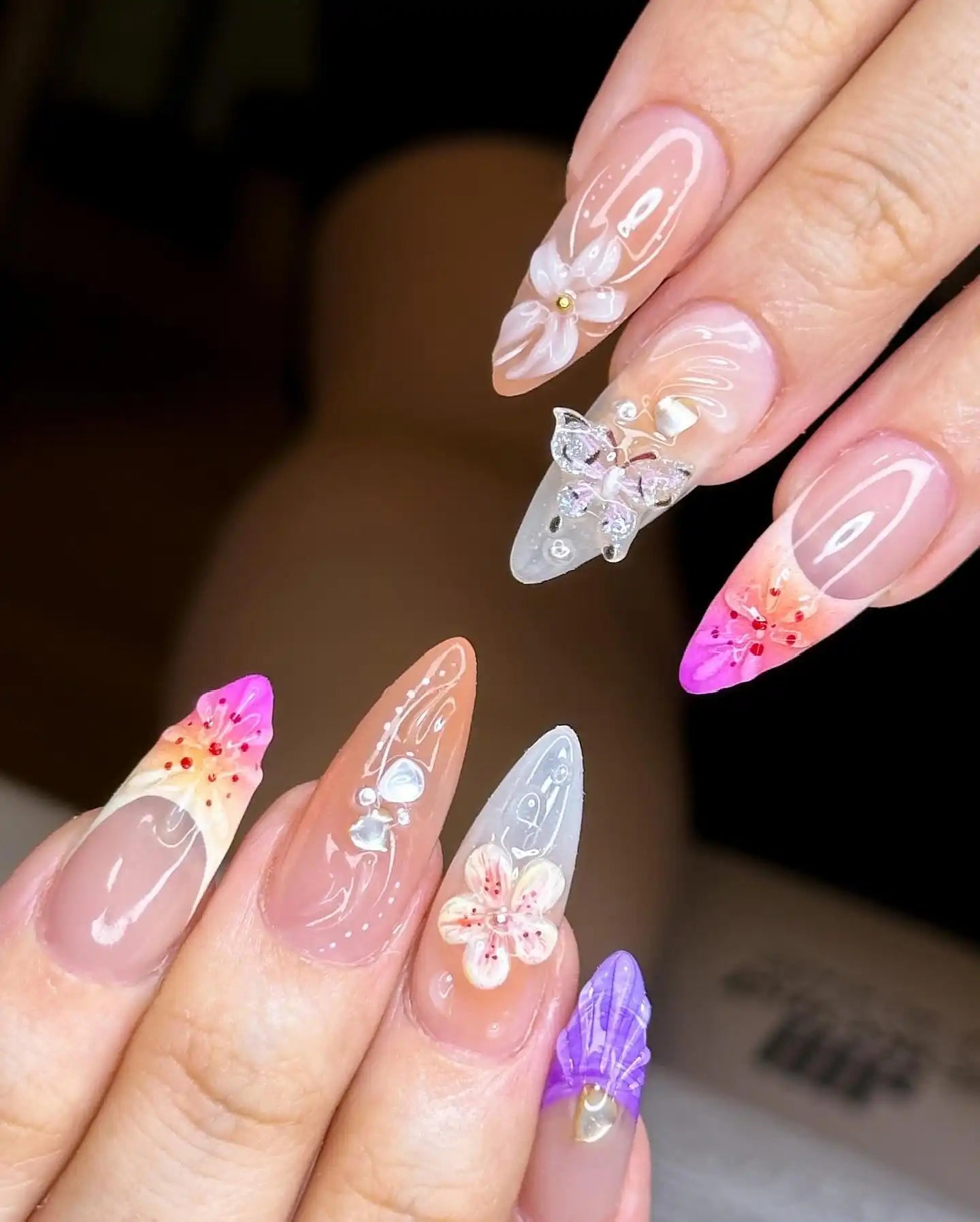 Hand with handmade press-on nails featuring 3D butterfly and flower designs, pastel colors, and gem accents.