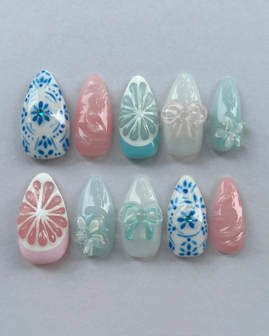 Handmade press-on nails with blue floral, pastel bow, and citrus designs on a gray background.