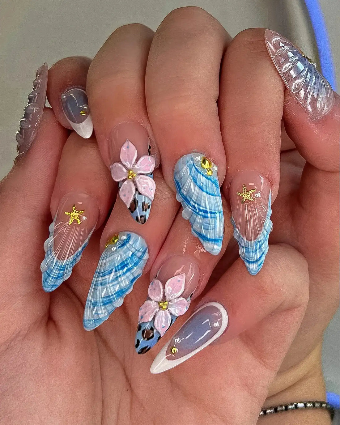 Hand with handmade press-on nails featuring blue seashell, pink flower, and gold starfish designs.