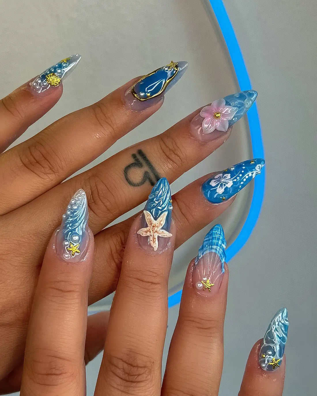 Hand with handmade press-on nails featuring blue seashell, pink flower, and gold starfish designs.