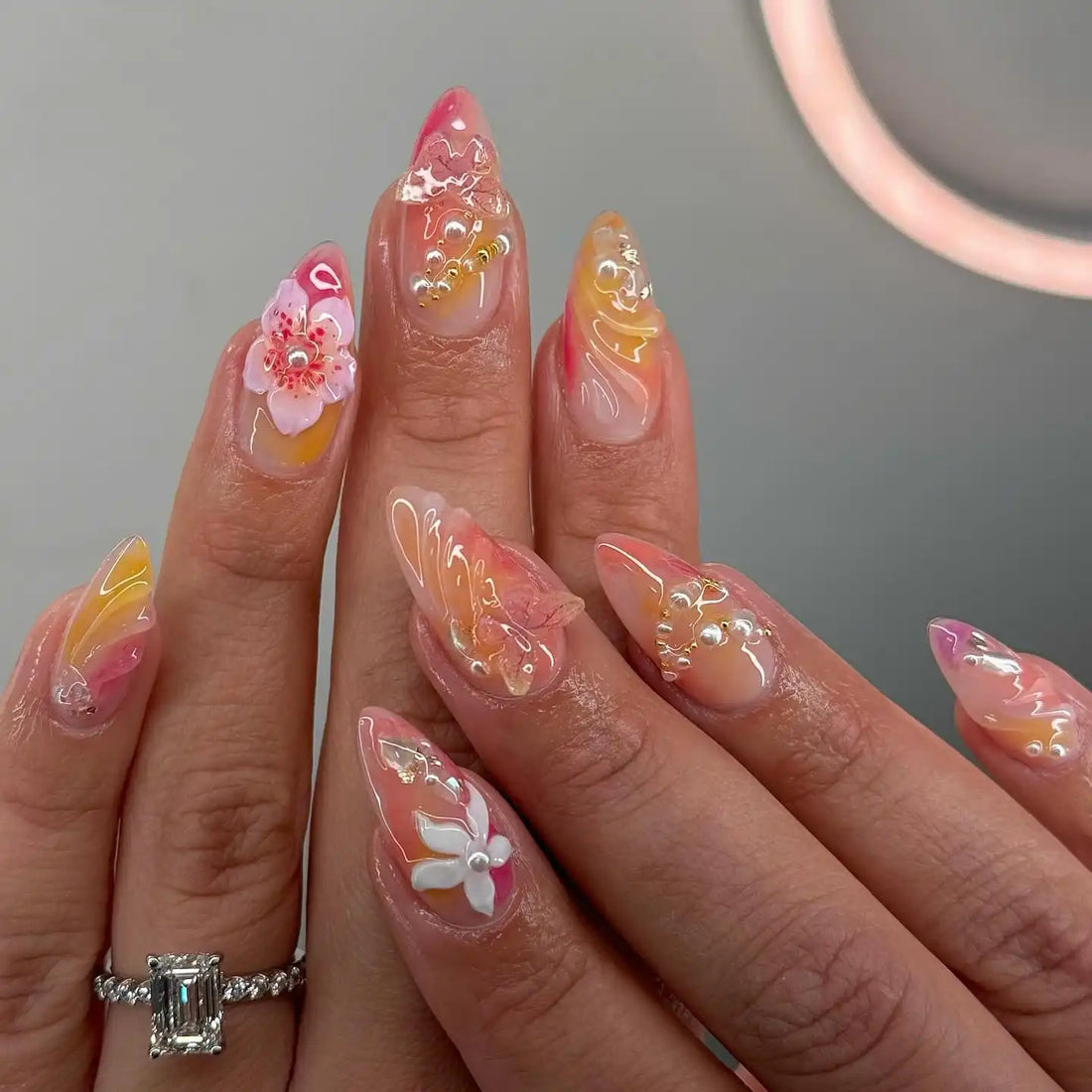 Hand with almond-shaped handmade press-on nails featuring floral, butterfly, and pearl designs in pink and yellow hues.