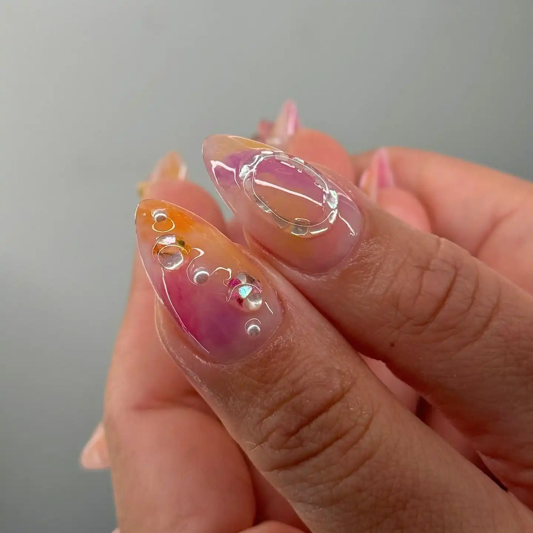 Hand with almond-shaped handmade press-on nails featuring floral, butterfly, and pearl designs in pink and yellow hues.