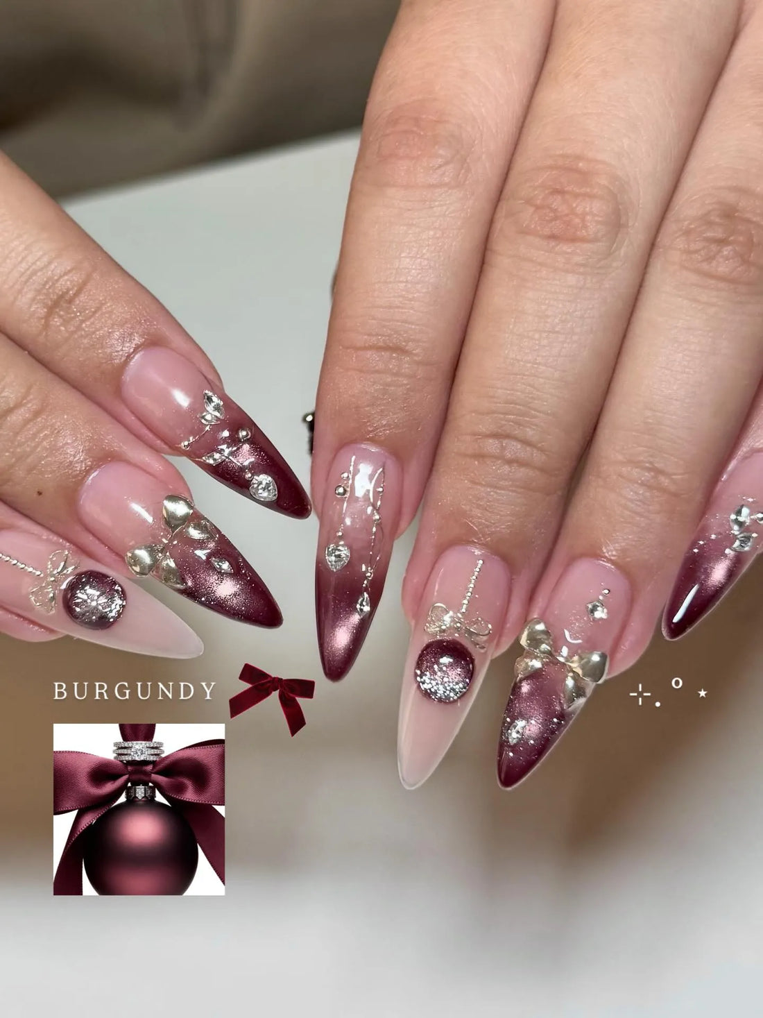 Hand with burgundy festive stiletto press-on nails, silver bows, rhinestones, and metallic accents