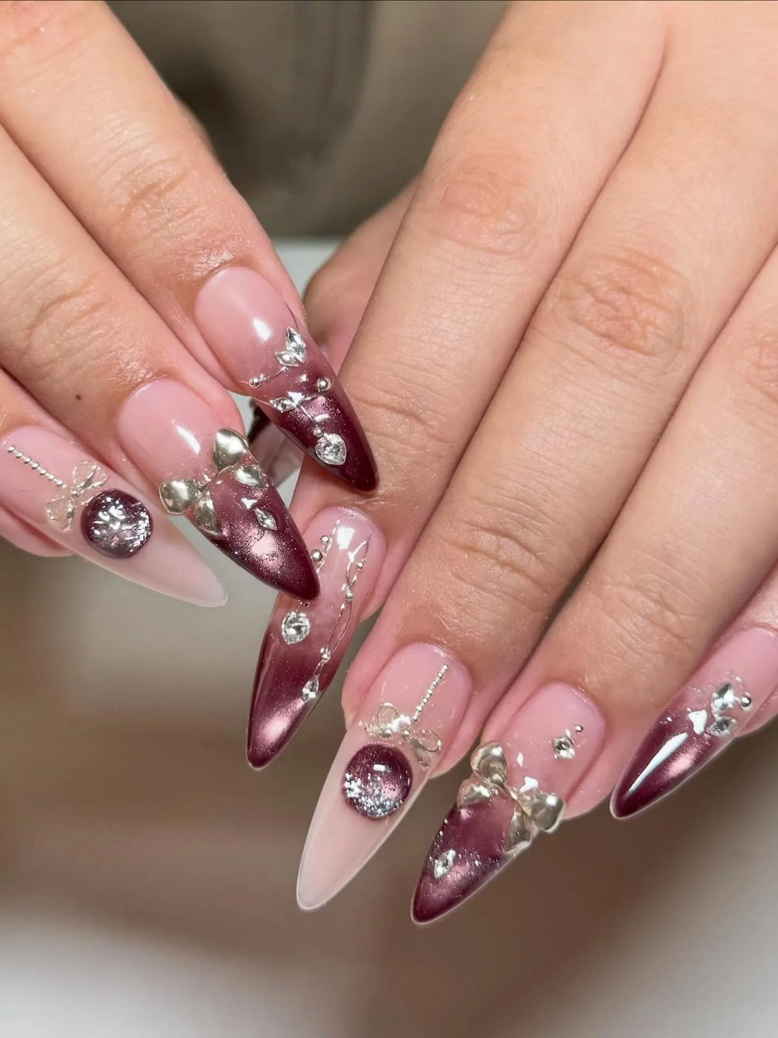 Hand with burgundy festive stiletto press-on nails, silver bows, rhinestones, and metallic accents