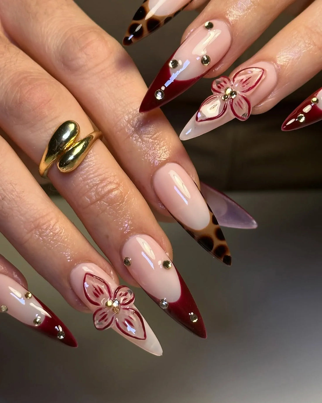 Hand with long stiletto press-on nails in burgundy red, leopard print, and 3D floral designs.