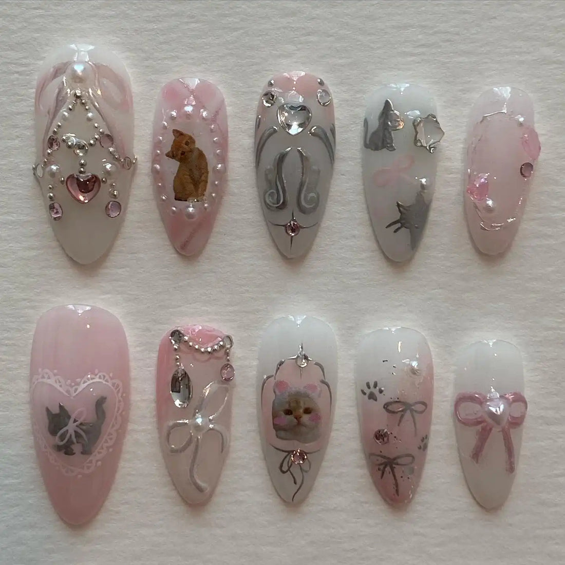 Handmade pink and white press-on nails with cat designs, pearls, bows, rhinestones, and lace accents.