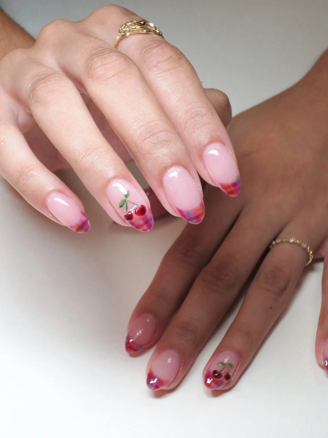 Hand with pink almond-shaped press-on nails featuring plaid French tips and cherry fruit nail art.