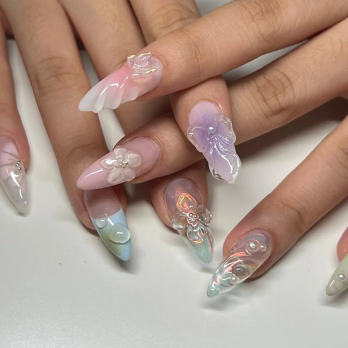 Hand with long handmade press-on nails featuring 3D floral and butterfly designs in pastel colors.