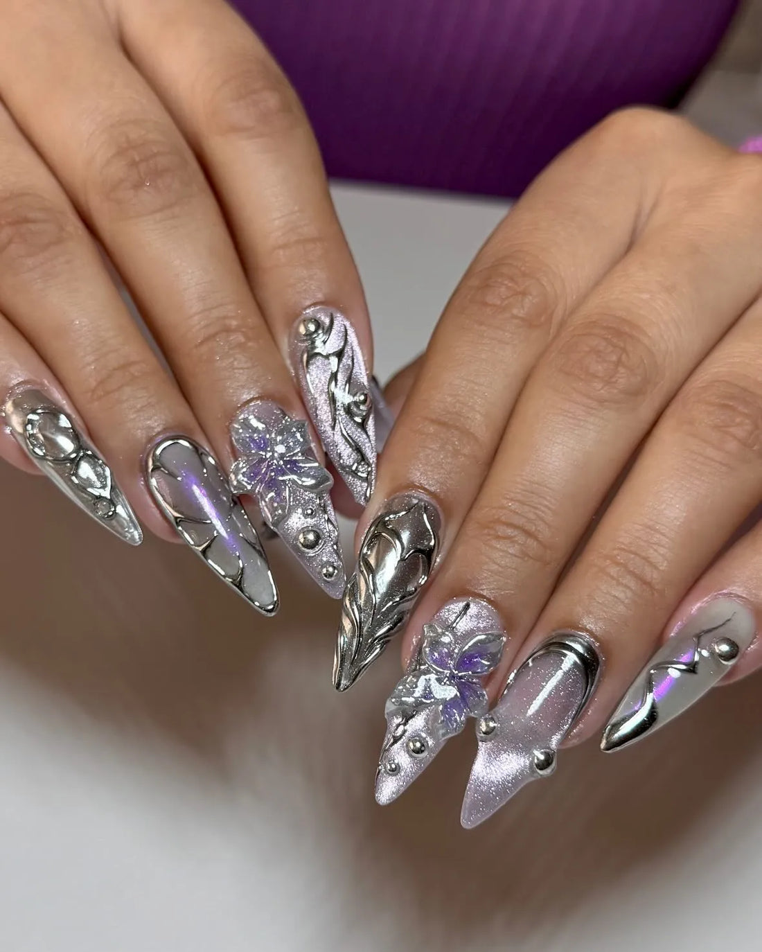 Hand with stiletto press-on nails in chrome and purple, featuring 3D metallic floral designs.