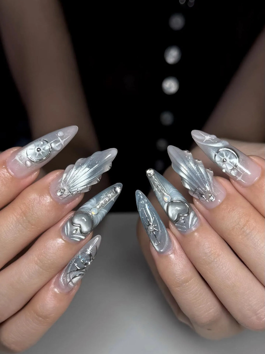 Hand with long stiletto silver press-on nails featuring heart and celestial 3D designs