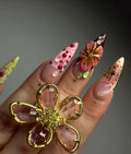 Hand with long stiletto press-on nails featuring 3D floral art, gems, and gold flower ring
