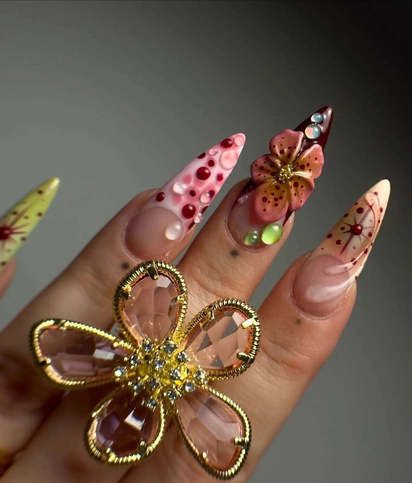 Hand with long stiletto press-on nails featuring 3D floral art, gems, and gold flower ring