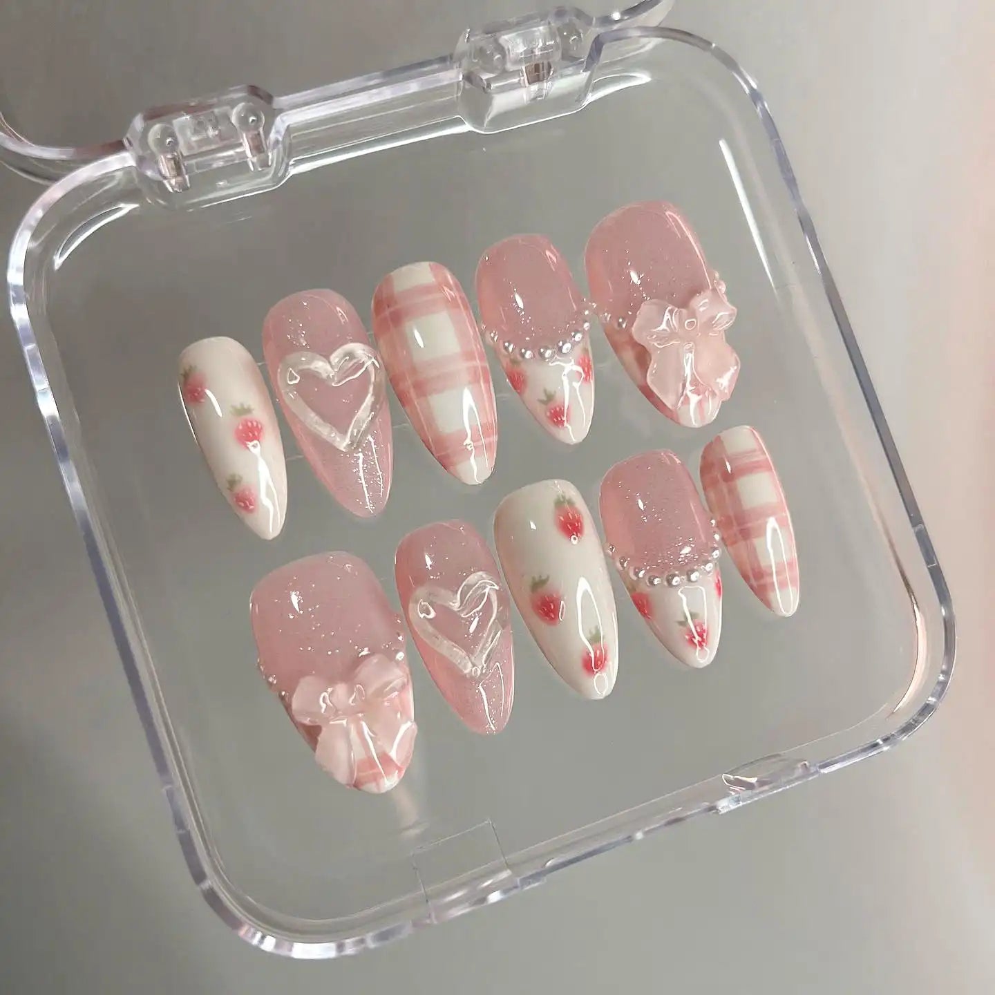 Handmade pink press-on nails with bows, hearts, pearls, plaid, and floral designs in a case
