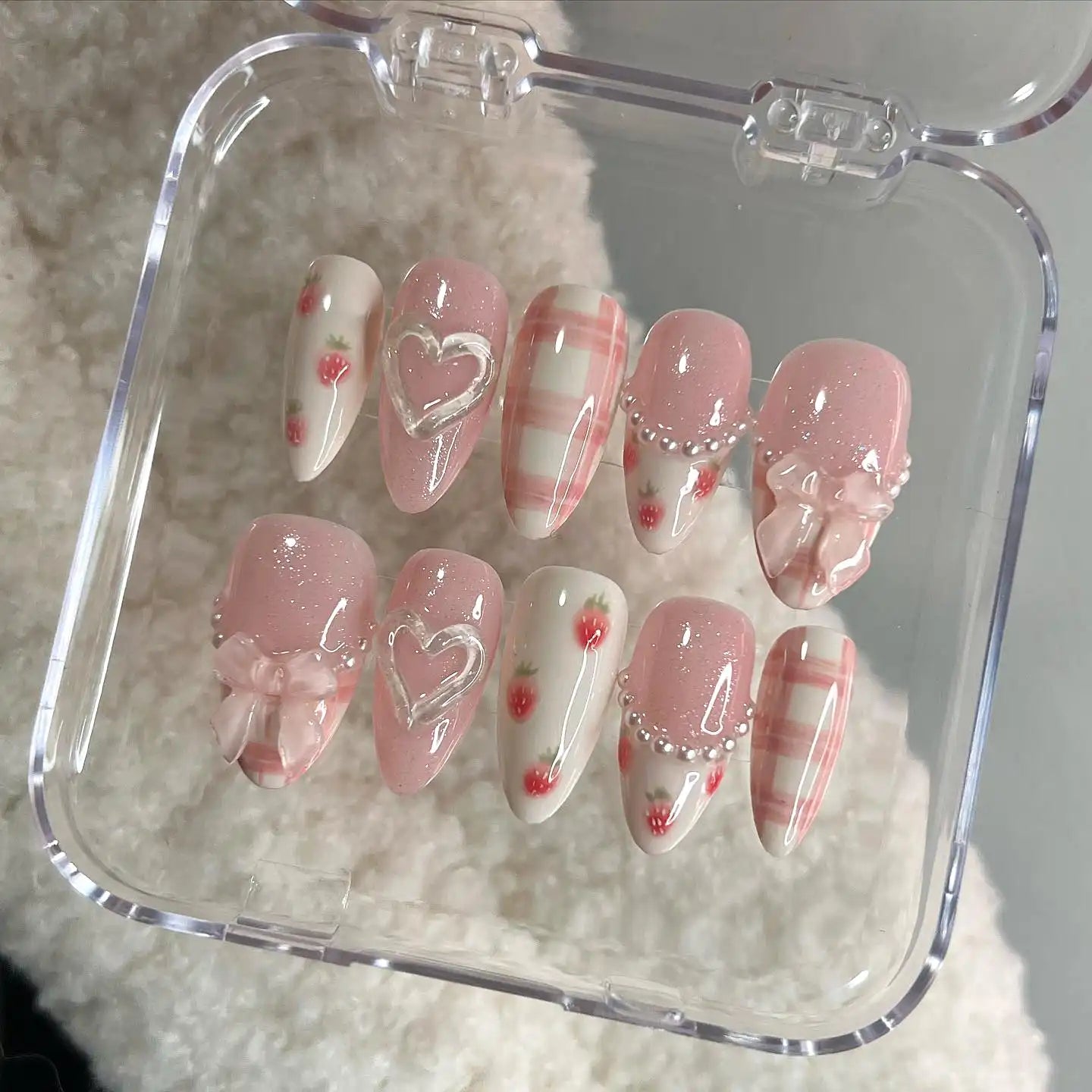 Handmade pink press-on nails with bows, hearts, strawberries, plaid and pearl accents in a case.