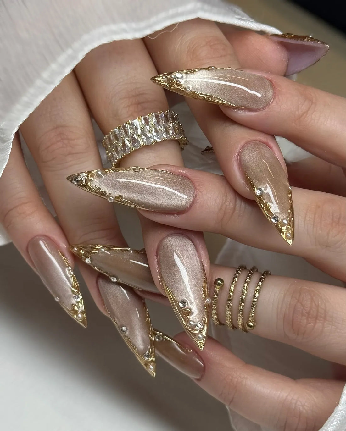 Hand with long stiletto press-on nails in metallic taupe, gold chrome tips, rhinestone accents.